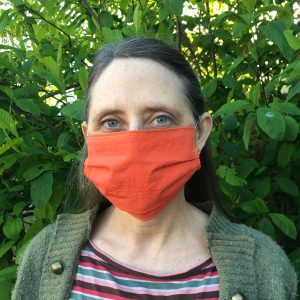woman wearing organic cotton orange poppy facemask with blue eyes and Indian plum leaves behind her. mask has elastic straps over the ears.