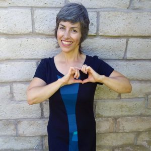 woman with short grey hair with hands in a heart shape and a smile. she is wearing a black and blue wearable art dress by vivid element.