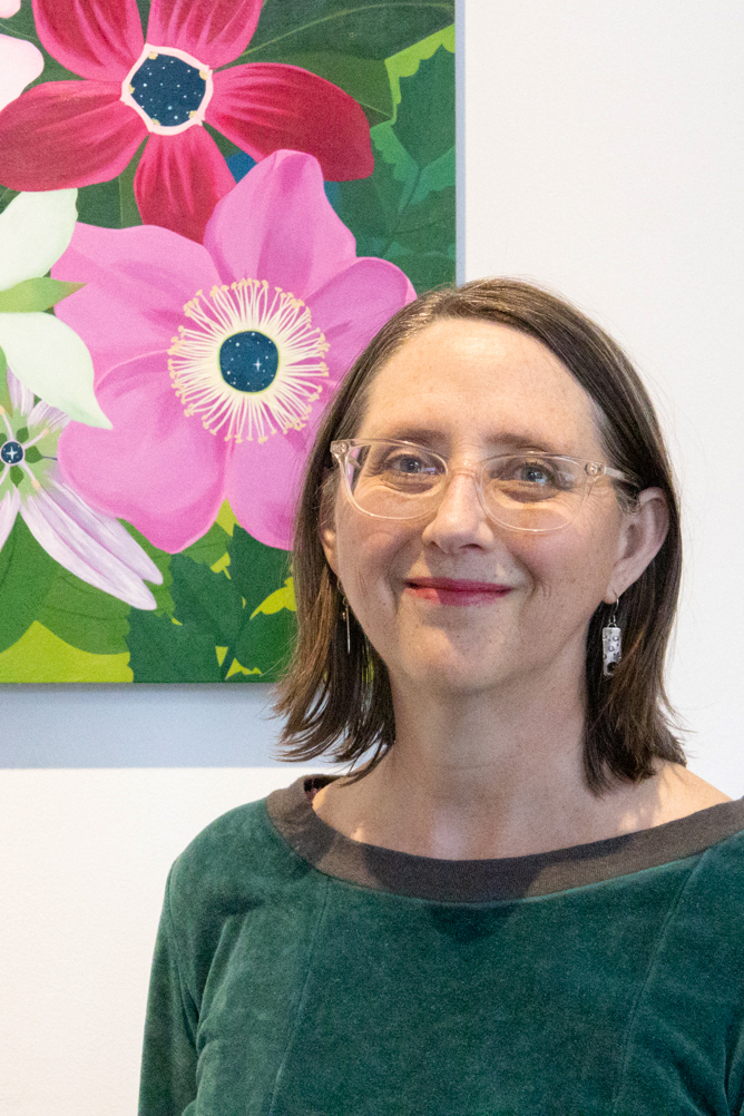 Artist Amy Daileda in front of one of her flower paintings. She is a white woman with clear glasses and a smile.